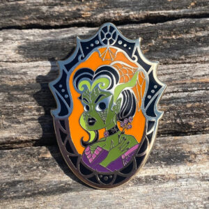 Olive green skinned Gothic Lady in a black Victorian frame enamel pin on a distressed wood background.