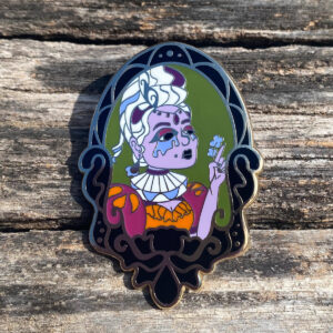 White haired and lavender complexed Gothic Lady in a black Victorian frame enamel pin on a distressed wood background.