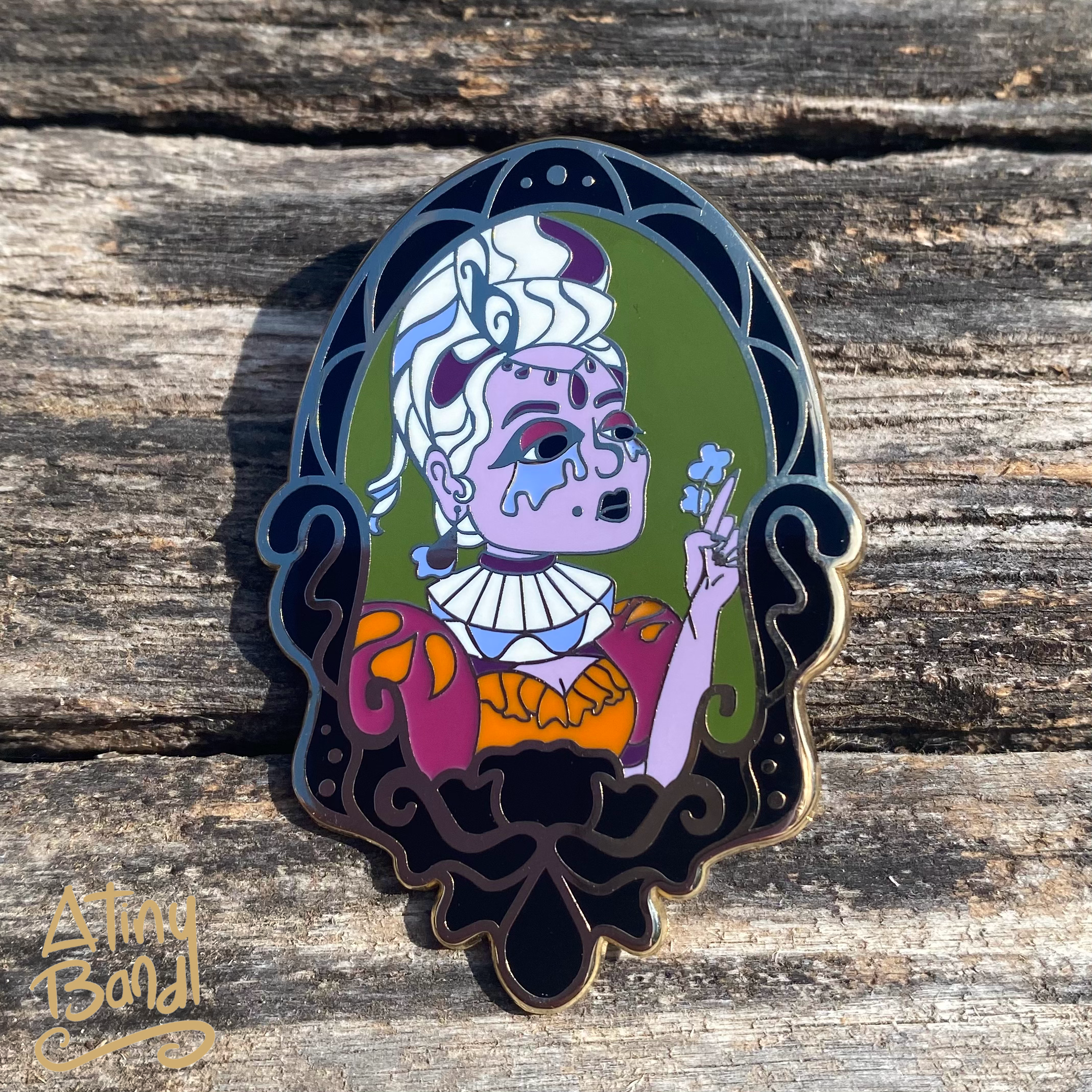 White haired and lavender complexed Gothic Lady in a black Victorian frame enamel pin on a distressed wood background.